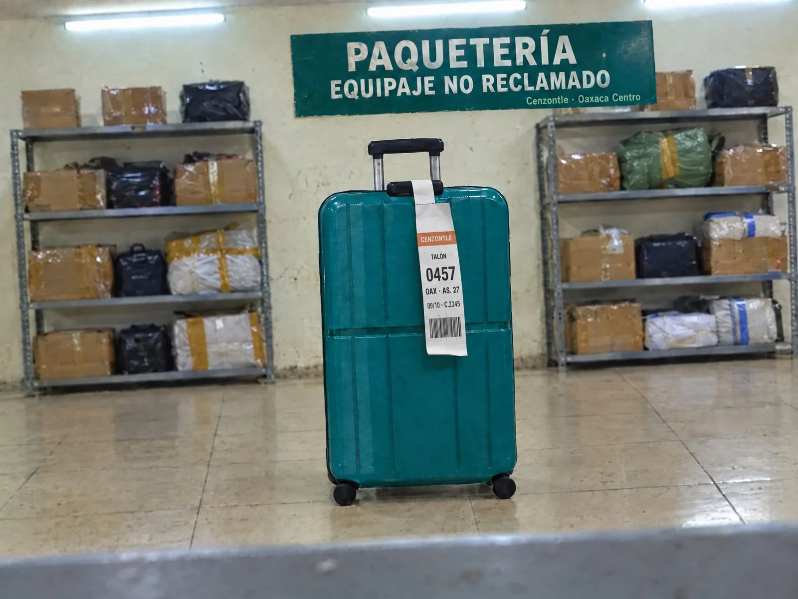 Una maleta verde azulado, sola, en la paquetería de la terminal de Oaxaca, bajo el letrero «Equipaje no reclamado».